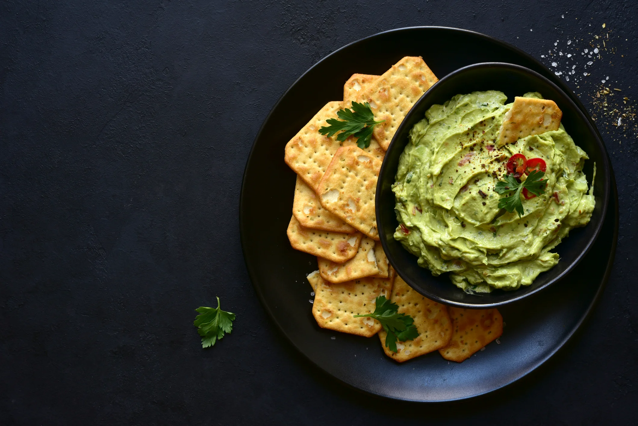 Avocado dip with crackers on black plate.